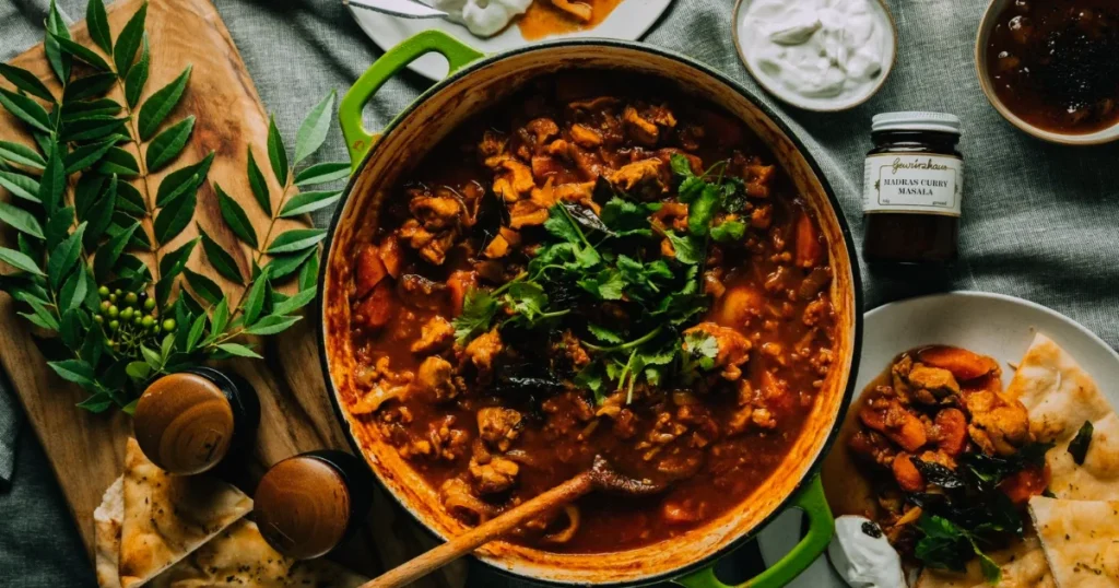 A pot of chicken Madras curry garnished with cilantro, served with naan, spices, and yogurt on a table.